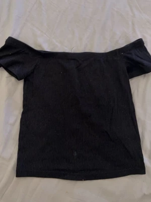 Svart offshoulder topp från Divided - Svart ribbad offshoulder topp från Divided. Toppens axlar är bara och den har en tight passform som framhäver figuren. Perfekt att styla med jeans eller kjol för en trendig look.