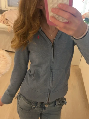 Blå zip-hoodie från Ralph Lauren - Snygg blå hoodie med dragkedja från Ralph Lauren. Klassisk passform med fickor framtill och liten rosa logga broderad på bröstet. Perfekt till jeans och enkel att matcha. Mjuk bomullskvalitet och ribbade muddar vid ärmslut och nederkant.