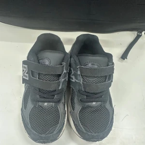 Svarta New Balance sneakers med kardborre 28 - Svarta sneakers från New Balance med meshpaneler och syntetdetaljer. Skorna har kardborreband för enkel påtagning och en vit sula med svarta och grå detaljer. Perfekta för en sportig och avslappnad stil.