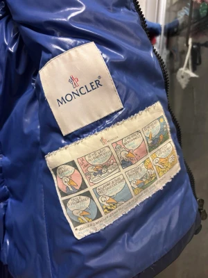 Blå dunjacka från Moncler - Snygg blå dunjacka från Moncler med glansig finish och klassisk quiltad design. Jackan har dragkedja framtill, två fickor med dragkedja och avtagbar huva. Insidan har Moncler-logga och ikonisk serietecknad etikett. Perfekt för kalla dagar. Fint skick men det finns ett litet hål i ena fickan, kan gå ner i pris vid snabb affär 🤗