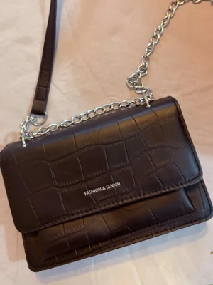 Svart axelväska från Fashion & Sennai - Snygg svart axelväska från Fashion & Sennai med croco-mönstrad yta och silverfärgad kedja. Väskan har ett stilrent och modernt utseende med lock och präglat mönster som ger extra edge till din outfit.