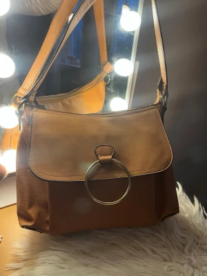 Brun handväska med metallring - Snygg brun handväska i skinn med axelrem och stor dekorativ metallring på framsidan. Väskan har en stilren design och praktisk storlek, perfekt för att bära dina viktigaste saker. Enkel att matcha med olika outfits och har en modern look.