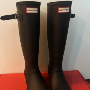 Svarta höga stövlar från Hunter - Klassiska svarta höga stövlar från Hunter med justerbart spänne vid skaftet och rund tå. Tillverkade i slitstarkt syntetmaterial som håller fötterna torra under regniga dagar. Perfekta för dig som vill ha både stil och funktion.