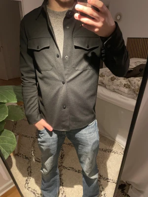 Svart overshirt från Zara (oanvänd) - Stilren svart overshirt från Zara Nypris 999❌ mitt pris 399✅ med klassisk krage och två bröstfickor med lock och knappar. Rak passform och långa ärmar, perfekt att slänga över en t-shirt eller hoodie. Tillverkad i ett mjukt material som känns skönt mot huden.
