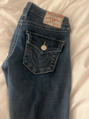 True Religion Joey Low Rise Flare Jeans - Snygga blå jeans från True Religion, modell Joey Low Rise Flare. Jeansen har låg midja, utställda ben och coola kontrastsömmar samt en ikonisk ficka med knapp bak. Perfekta för dig som gillar en retro och avslappnad look. Uppsyda i längden