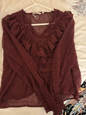 Vinröd blus med volanger och spets - Vinröd blus i transparent tyg med diskret rutmönster, volangdetaljer längs V-ringningen och spetsinslag. Långa ärmar med breda smockade muddar ger en romantisk look. Perfekt för dig som gillar feminina detaljer och trendig stil. Den är från only, köpt för 500 kr 