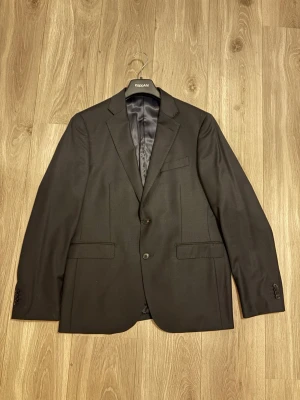 Svart kavaj från these glory days - Stilren svart kavaj från these glory days med klassisk passform. Kavajen har två knappar framtill, fyra dekorativa knappar vid ärmsluten och tre fickor på framsidan. Tillverkad i ett slätt tyg med diskret glans och helfodrad insida. Helt ny utan prislapp endast testad en gång inget är uppsprätt