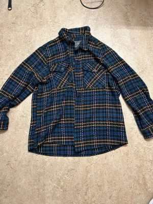 Rutig overshirt från Primark - Overshirt från Primark i blått, svart och grönt med rutigt mönster. Klassisk skjortkrage, två bröstfickor med knappar och långa ärmar. Perfekt lager på lager-plagg för dig som gillar avslappnad stil.
