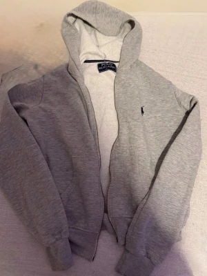 Grå hoodie från Polo Ralph Lauren - Snygg grå hoodie från Polo Ralph Lauren med dragkedja och klassisk huva. Tröjan har det ikoniska mörkblå polospelar-loggan broderad på bröstet och ribbade muddar vid ärmar och nederkant. Tillverkad i mjukt sweatshirtmaterial för extra komfort. (Dragkedjan är trasig )