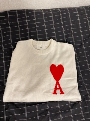 Vit stickad tröja från Ami med rött hjärta - Vit stickad tröja från Ami med ett stort rött hjärta och bokstaven A på bröstet. Tröjan har rund hals, långa ärmar och breda ribbade muddar vid ärmslut och nederkant. Perfekt för dig som gillar stilrena plagg med en twist. Bara testad på annars  i nyskick, inga defekter. Hör av dig om du har några frågor.:) behöver få såldt snabbt så kan gå ner till 500 vid snabb affär