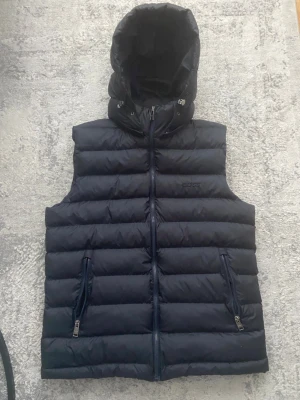 Svart dunväst från BOSS med huva - Snygg svart dunväst från BOSS med avtagbar huva och två dragkedjeförsedda fickor. Västen har quiltad design och diskret BOSS-logga på bröstet. Perfekt för lager-på-lager och håller dig varm under kyliga dagar.