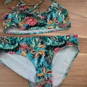 Färgglad bikini med djungelmönster storlek 146/152 - Säljer en bikini med tropiskt djungelmönster i grönt, rosa, gult och blått. Överdelen har korsade axelband och bygel, underdelen är klassisk brief-modell. Mönstret har blommor och leoparder för en cool och unik look. Perfekt för stranden eller poolen!