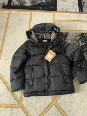 Svart dunjacka med huva från  berbery  - Snygg svart dunjacka från Lexington med avtagbar huva och rutigt innerfoder. Jackan har dragkedja och knappar framtill samt två stora fickor. Perfekt för kalla dagar och har en klassisk pufferlook.