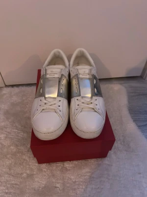Valentino Open sneakers vit/silver - Snygga Valentino Open sneakers i vitt skinn med bred silverfärgad detalj över mitten. Klassisk rund tå, platt sula och coola nitar på hälen. Perfekta för dig som vill ha en stilren men ändå unik look.skriv vid frågor!