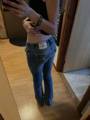 True Religion Becca Bootcut Jeans - Säljer ett par blå bootcut jeans från True Religion, modell Becca. oklassiska fickor med lock och gul kontrastsöm, samt snygga silverfärgade knappar. Jeansen är i stretchigt denim och har en ikonisk patch bak.