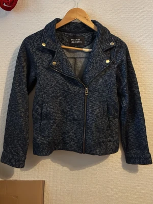 Mörkblå bomberjacka Galeries Lafayette - Snygg mörkblå bomberjacka från Galeries Lafayette med mönstrad yta och guldfärgade knappar. Jackan har dragkedja framtill, långa ärmar och en cool krage. Materialet är mjukt och känns som bomull, perfekt för dig som vill ha en chill och stilren look.