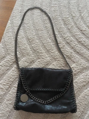 Svart axelväska med kedjedetaljer - Cool svart axelväska med grov silverfärgad kedja som axelrem och runt kanterna. Väskan har ett lock med magnetknapp och insidan är fodrad med leopardmönster. Materialet ser ut som skinn och väskan har en rund metallbricka som detalj.