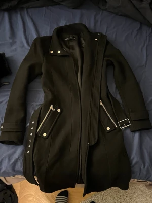 Svart kappa med bälte från Zara Woman - Snygg svart kappa från Zara Woman med markerad midja och bälte med metallspänne. Jackan har långa ärmar, dragkedjor vid fickorna och dekorativa knappar. Materialet är ullblandning och modellen är figurnära med coola detaljer.
