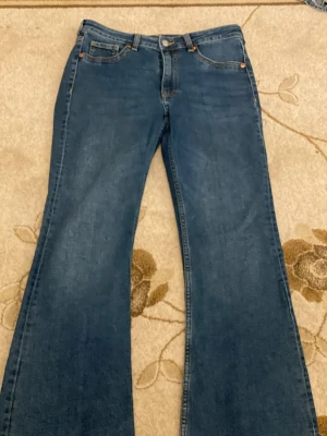 Blå bootcut jeans i storlek 42 - Säljer ett par klassiska blå bootcut jeans i storlek 42. Jeansen har fem fickor, knapp och dragkedja framtill samt bälteshällor. Materialet är slitstarkt jeans och passformen är normal med snyggt utsvängda ben. Perfekt för dig som gillar retrovibbar och vill ha en cool siluett.