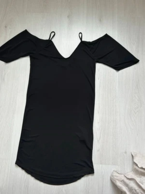 Svart offshoulder kortklänning - Snygg svart kortklänning med offshoulder och smala axelband. Klänningen har en v-ringad hals och är tillverkad i ett mjukt, stretchigt material som ger en skön passform. Perfekt för dig som gillar stilrena och enkla plagg med en modern touch.