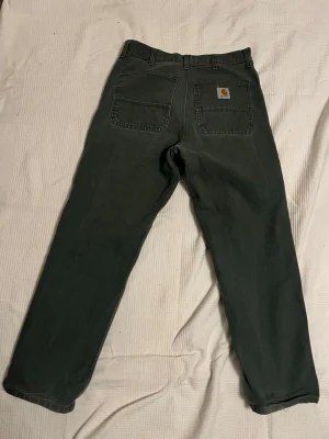 Mörkgröna Carhartt byxor med raka ben - Säljer ett par mörkgröna Carhartt byxor med raka ben och klassisk passform. Byxorna har två stora bakfickor, varav en med Carhartt-logga, samt robust knapp och dragkedja framtill. Materialet är slitstark bomull, perfekt för dig som gillar streetwear.