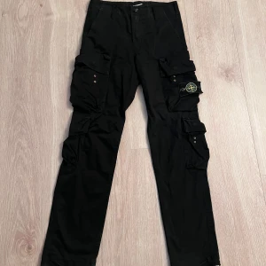 Svarta Stone Island cargopants  - Svarta cargopants från Stone Island med flera fickor på benen och klassisk logopatch på vänster sida. Byxorna har en rak passform och är tillverkade i bomull. Perfekt för dig som gillar streetwear och funktionella detaljer. Byxorna gör lite ljud när man går dock