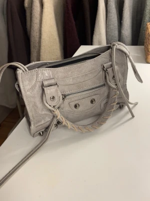 Grå handväska  - Säljer en ljusgrå handväska med silvriga detaljer och dragkedja. Med väskan tillkommer även ett långt band som syns lite på sista bilden. Skriv för bättre bilder, frågor eller mått!