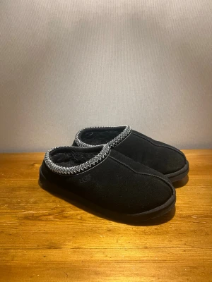 UGG Tasman - Orginalpris: 1699kr. Knappt använda!Svarta skor från UGG med mjuk mocka på utsidan och fluffigt foder. Snyggt mönstrad kant i vitt och svart runt öppningen. Platt sula med grovt mönster för extra grepp. Perfekt för chill dagar hemma eller när du vill ha det extra bekvämt. 