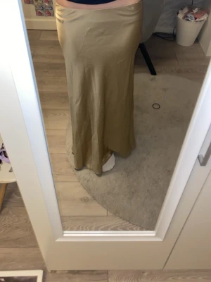 Guldig långkjol i satin - Säljer en snygg guldig långkjol med glansig satinfinish. Kjolen har en rak och flowy siluett som sitter högt i midjan och faller mjukt ner mot golvet. Perfekt för dig som gillar stilrena och eleganta plagg med lite extra lyxig känsla.