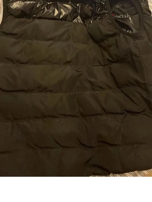 Svart dunväst från Moncler - Säljer en svart dunväst från Moncler med quiltad design och glansiga detaljer upptill. Västen har ett klassiskt snitt och är tillverkad i polyester med dunfyllning. Insidan har Moncler-loggor tryckta över hela fodret för en lyxig känsla. Perfekt för kalla dagar.