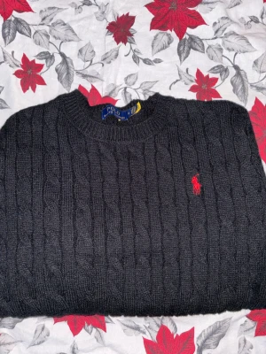 Svart stickad tröja Polo Ralph Lauren - Snygg svart stickad tröja från Polo Ralph Lauren i storlek M. Tröjan har kabelstickat mönster, rund hals och lång ärm. På bröstet finns den klassiska röda Polo-loggan broderad. Perfekt för dig som gillar stilrena och tidlösa plagg.