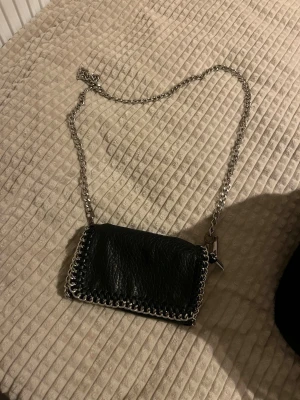 Svart axelväska med kedja från Tiamo - Snygg svart axelväska från Tiamo i skinn med grov silverkedja som axelrem. Väskan har ett rektangulärt format och detaljer med kedja runt kanterna. Insidan har dragkedja och fack för småsaker. Perfekt för att lyfta din outfit.