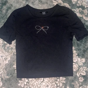 Svart croptop med rosettdetalj från House - Svart kortärmad topp från House i bomull med en glittrig rosettdetalj framtill. Rund halsringning med söt spetskant och enkel, croppad passform. Perfekt för dig som gillar stilrena plagg med en liten twist. Använd en eller två gånger. 