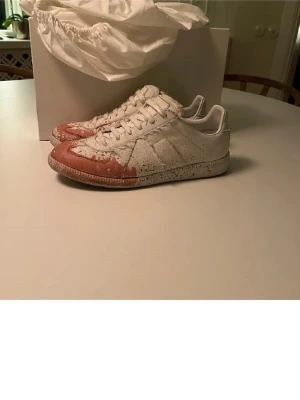 Maison Margiela sneakers med färgstänk - Maison Margiela sneakers i vitt med unika röda och beige färgstänk över tåpartiet. Klassisk låg modell med snörning och rund tå. Tillverkade i skinn och har en cool, konstnärlig vibe som sticker ut. Perfekta för dig som vill ha något annorlunda i garderoben. Solklart äkta!