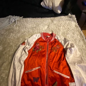 Röd bomberjacka med broderade detaljer - Snygg röd bomberjacka med vita ärmar och broderade rosor och fjärilar både fram och bak. Jackan har ribbade muddar, dragkedja och coola broderier.