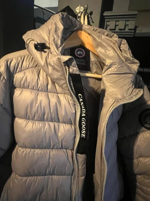 Ljusgrå pufferjacka från Canada Goose - Säljer en snygg ljusgrå pufferjacka från Canada Goose med huva och broderad logga på ärmen. Jackan har dragkedja framtill och är quiltad för extra värme. Perfekt för kalla dagar och har en clean look med diskreta detaljer.