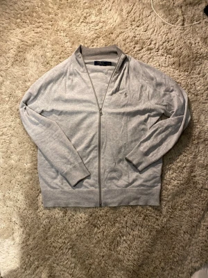Grå kofta från Polo Ralph Lauren - Säljer en stilren grå kofta från Polo Ralph Lauren med dragkedja framtill och diskret logga på bröstet. Tröjan har lång ärm, ribbade muddar och är gjord i mjuk bomull. Perfekt till lager på lager under höst och vår.