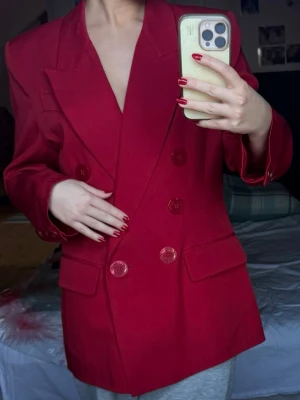 Röd dubbelknäppt kavaj - Säljer en snygg röd kavaj med dubbelknäppning och breda slag. Kavajen har stora matchande knappar och två fickor framtill. Passformen är så snygg och materialet känns som bomull eller polyester. Perfekt för dig som vill sticka ut med färg. Vaddering i axlarna.