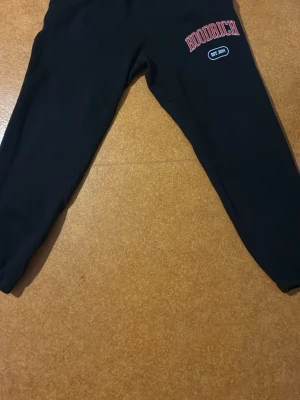 Svarta Hoodrich mjukisbyxor - Svarta mjukisbyxor från Hoodrich med tryck i vitt och rött på benet och bakfickan. Byxorna har elastiska muddar vid bensluten och en normal passform. Materialet är mjukt och bekvämt, perfekt för chill eller streetwear. Snygga detaljer med broderad logga och text.