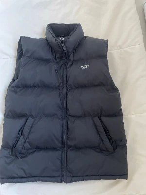 Svart dunväst från WESC - Säljer en svart dunväst från WESC med hög krage och två fickor med dragkedja. Västen har quiltad design och stängs med dragkedja framtill. Perfekt att slänga över en hoodie när det är kyligt ute.