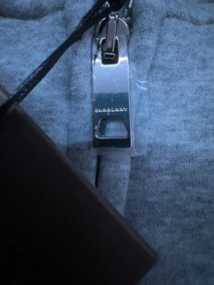 Grå zip hoodie från Burberry - Grå zip hoodie från Burberry med broderad ryttare på bröstet och klassisk dragkedja med Burberry-logga. Hoodien har mjukt material och ribbade muddar. Insidan har en beige rutig detalj vid kragen. Perfekt för dig som vill ha en stilren och lyxig look.