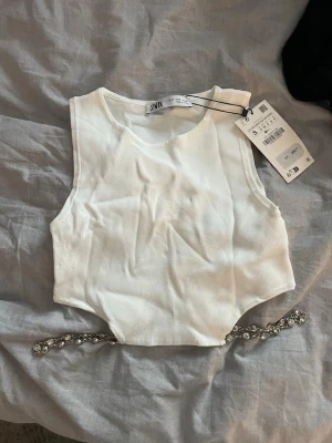 Vit croppad linnetopp från Zara - Säljer en vit croppad linnetopp från Zara i storlek S. Toppen är ärmlös och har en cool detalj med en glittrig kedja längst ner. Materialet är ribbad och känns mjukt mot huden. Perfekt för att lyfta din outfit med en snygg accessoar.