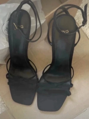 Svarta klackade sandaler från Mango - Säljer ett par svarta sandaler från Mango med smala remmar och snygg rosettdetalj framtill. Skorna har fyrkantig tå, hög klack och ankelsnörning som ger en trendig look. Perfekta för dig som gillar stilrena och moderna skor.