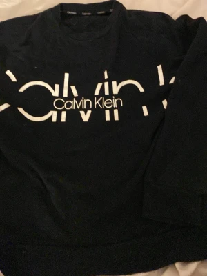 Svart sweatshirt från Calvin Klein - Svart sweatshirt från Calvin Klein med stor vit logotyp framtill. Tröjan har rund hals och långa ärmar, perfekt för en chill och stilren look. Materialet är mjukt och skönt, troligen bomull eller polyester. Passar dig som gillar klassiska märken och enkel design.