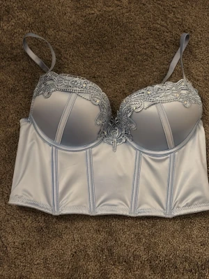 Ljusblå korsett med spetsdetaljer - Säljer en ljusblå korsett med vackra spetsdetaljer och små pärlor på kuporna. Toppen har justerbara axelband och flera hyskknäppningar i ryggen för perfekt passform. Materialet är glansigt och känns lyxigt mot huden. Perfekt för dig som vill ha något extra fint. Prislappen kvar 