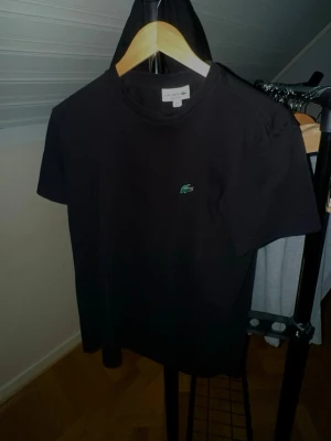 Svart t-shirt från Lacoste - Svart t-shirt från Lacoste med klassisk krokodillogga broderad på bröstet. Regular fit och rund hals. Materialet är mjuk bomull som känns skönt mot huden. Perfekt för dig som gillar stilrena och enkla plagg med en ikonisk touch.