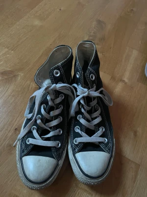 Svarta Converse (höga) - Säljer ett par klassiska höga svarta Converse med vit gummitå och vita snören. Skorna har rund tå, platt sula och tydliga vita sömmar. Märket syns på sidan.