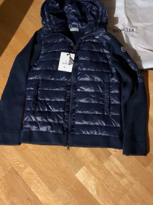 Mörkblå pufferjacka från Moncler - Snygg mörkblå pufferjacka från Moncler med huva och stickade ärmar. Jackan har quiltad framsida, dragkedja, praktiska fickor och Moncler-logga på ärmen. bekvämt. Skriv om ni har frågor. Storlek L. 