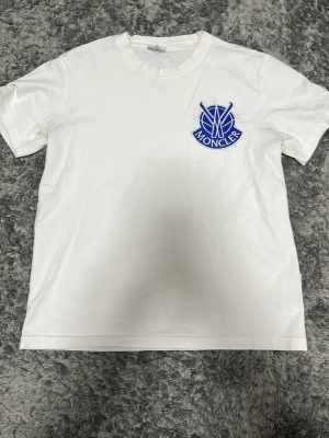 Vit Moncler t-shirt med blå logga - Säljer en fräsch vit t-shirt från Moncler med en stor blå broderad logga på bröstet. T-shirten har rund hals och korta ärmar, perfekt för dig som gillar stilrena och exklusiva plagg. Materialet är mjuk bomull som känns skönt mot huden.