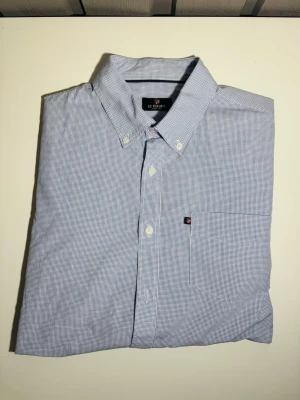 Blårutig skjorta från U.S. Polo Assn - Snygg blårutig skjorta från U.S. Polo Assn med klassisk krage och knappar framtill. Skjortan har en bröstficka med liten logotyp och är långärmad. Materialet är mjuk bomull och passformen är normal. Perfekt för dig som gillar stilrena och tidlösa plagg.